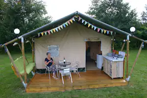 Zelt mit Holzpodest, Campingmöbeln und Küchenzeile im Grünen, umgeben von Bäumen und Rasen.
