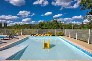 Camping-Pool mit gelber Rutsche, Liegen am Beckenrand und Zaun, im Hintergrund Bäume und blauer Himmel mit Wolken.