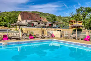 Schwimmbad mit blauem Wasser, Liegestühlen und Sonnenschirmen vor einem Ferienhaus mit Reetdach und Terrasse.