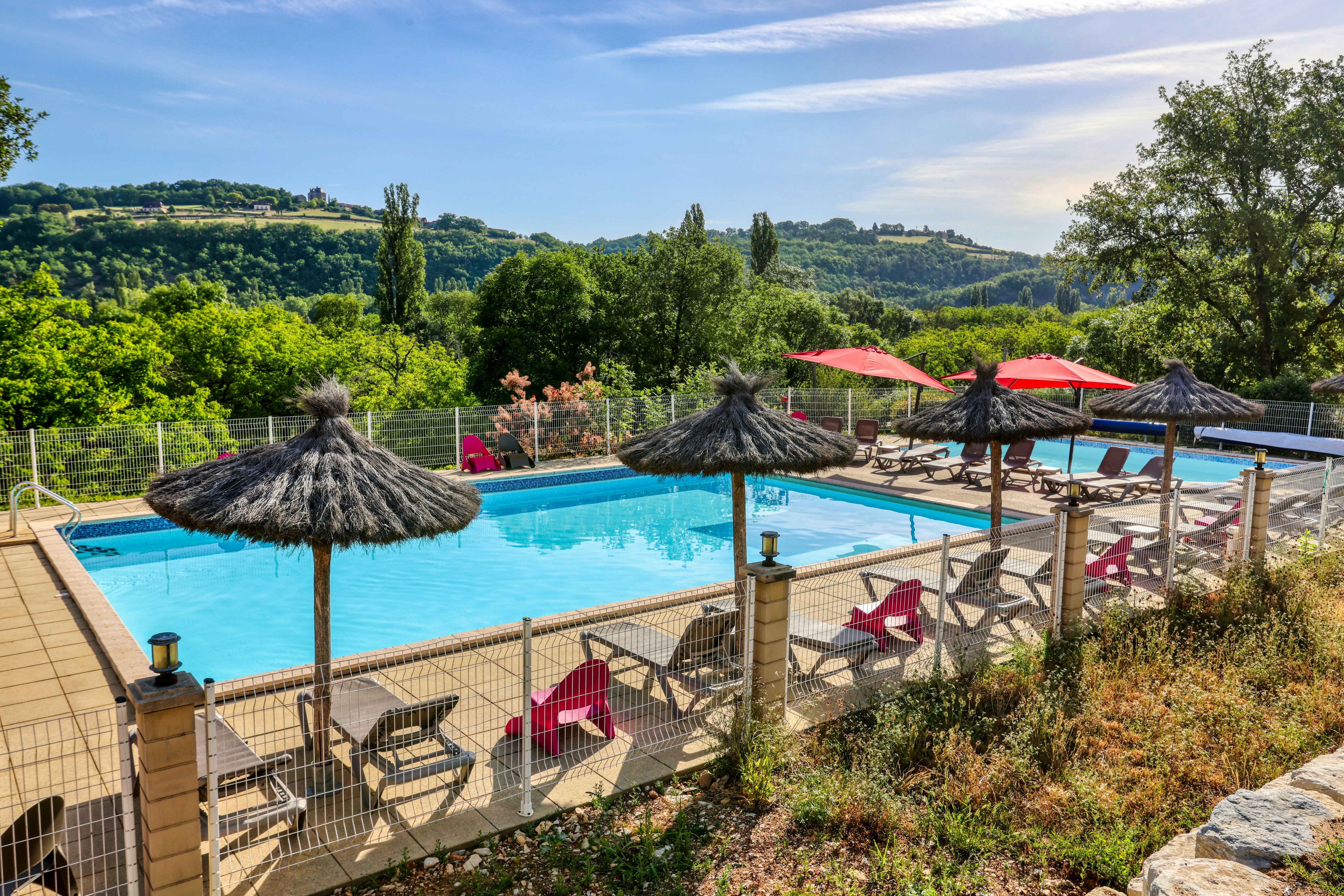 Schwimmbad mit Liegen und Sonnenschirmen auf einer Terrasse, dahinter grüne Hügel und Bäume unter klarem Himmel.