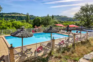Schwimmbad mit Liegen und Sonnenschirmen auf einer Terrasse, dahinter grüne Hügel und Bäume unter klarem Himmel.