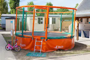 Oranges Trampolin auf dem Campingplatz mit Leiter, daneben ein kleines Laufrad und Gebäude im Hintergrund.