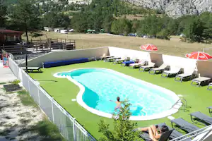 Großer Campingpool mit Liegen und Sonnenschirmen auf einer Rasenfläche, umzäunt mit Blick auf Bäume und Felsen.