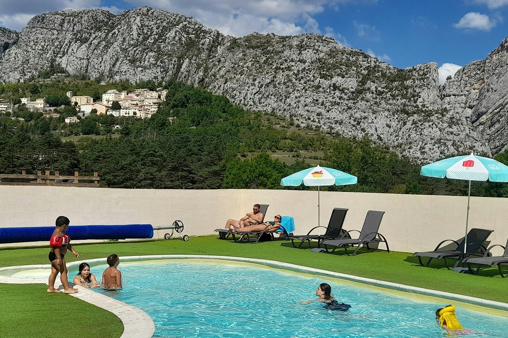 Camping-Pool mit Liegen und Sonnenschirmen, Kinder im Wasser, im Hintergrund Berge und Felswände.