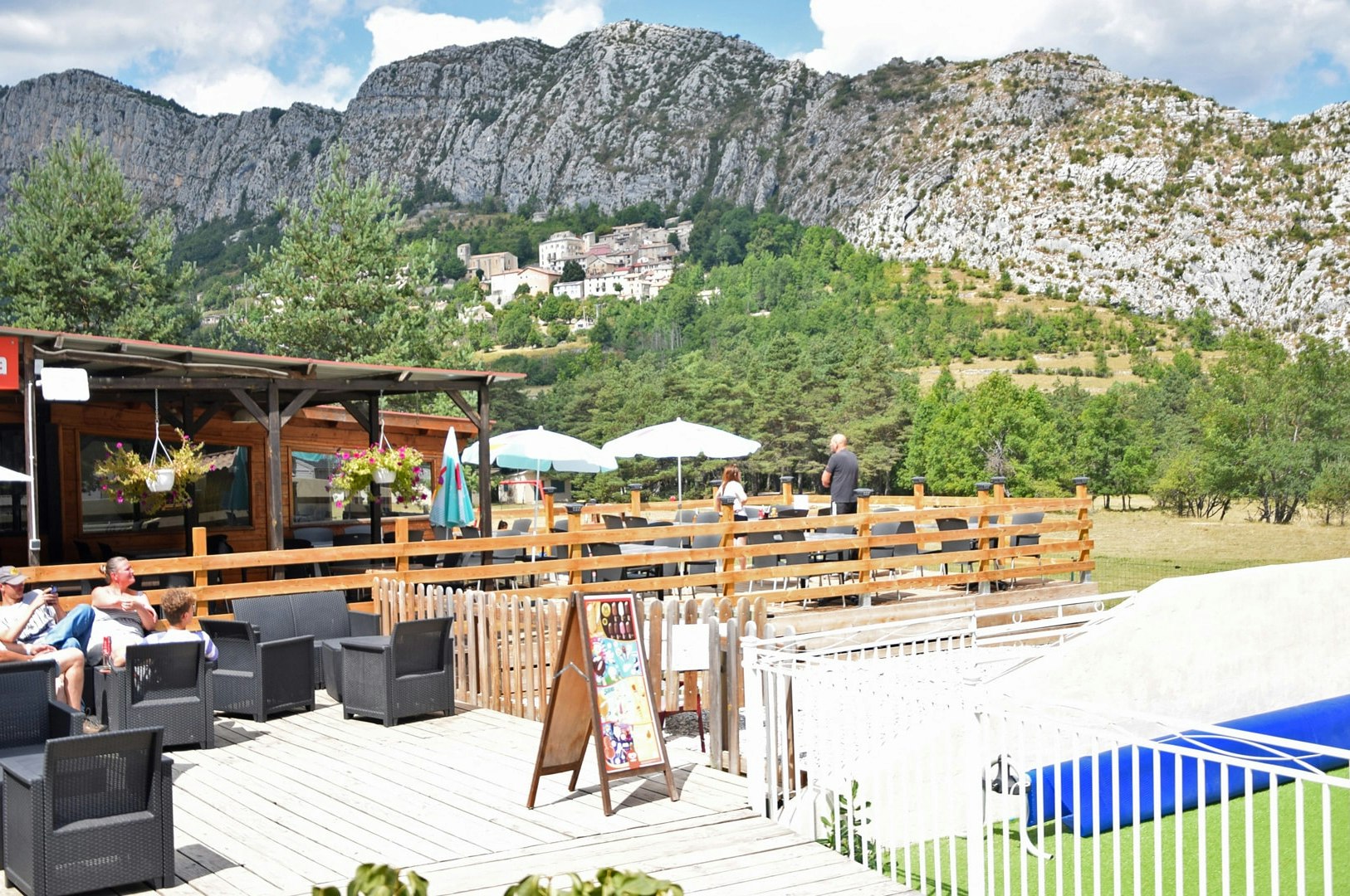 Camping-Terrasse mit Holzzaun, Sitzgruppen und Sonnenschirmen, im Hintergrund Berge und grünes Tal.