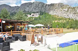 Camping-Terrasse mit Holzzaun, Sitzgruppen und Sonnenschirmen, im Hintergrund Berge und grünes Tal.