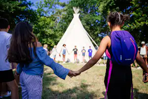 Zelt im Grünen, Kinder und Erwachsene laufen über eine Wiese, im Hintergrund weitere Zelte im Campingbereich.