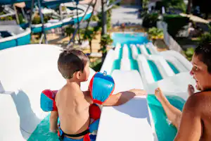 Ein Kind mit Schwimmflügeln und eine Person am Rand eines Camping-Schwimmbeckens, im Hintergrund weitere Liegen und Pflanzen.