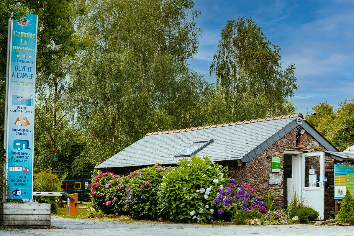 Kleines Campinggebäude mit weißer Tür und Fenster, Schrägdach und Blumenbeet vor dem Eingang, umgeben von Bäumen.