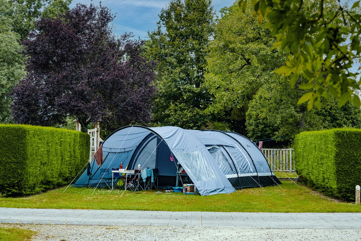 Zelt im Campingplatz mit zwei Eingängen, davor Gartenmöbel und ein kleiner Tisch, umgeben von Hecken und Bäumen.