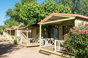 Bungalow mit Veranda, Holztreppe und Blumenbeeten vor grünen Bäumen, im Hintergrund weitere Bungalows.