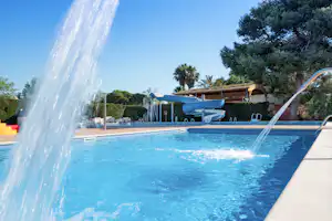 Schwimmbecken mit Wasserrutsche, Sprühfontänen und Liegen am Rand, umgeben von Hecken und Bäumen.