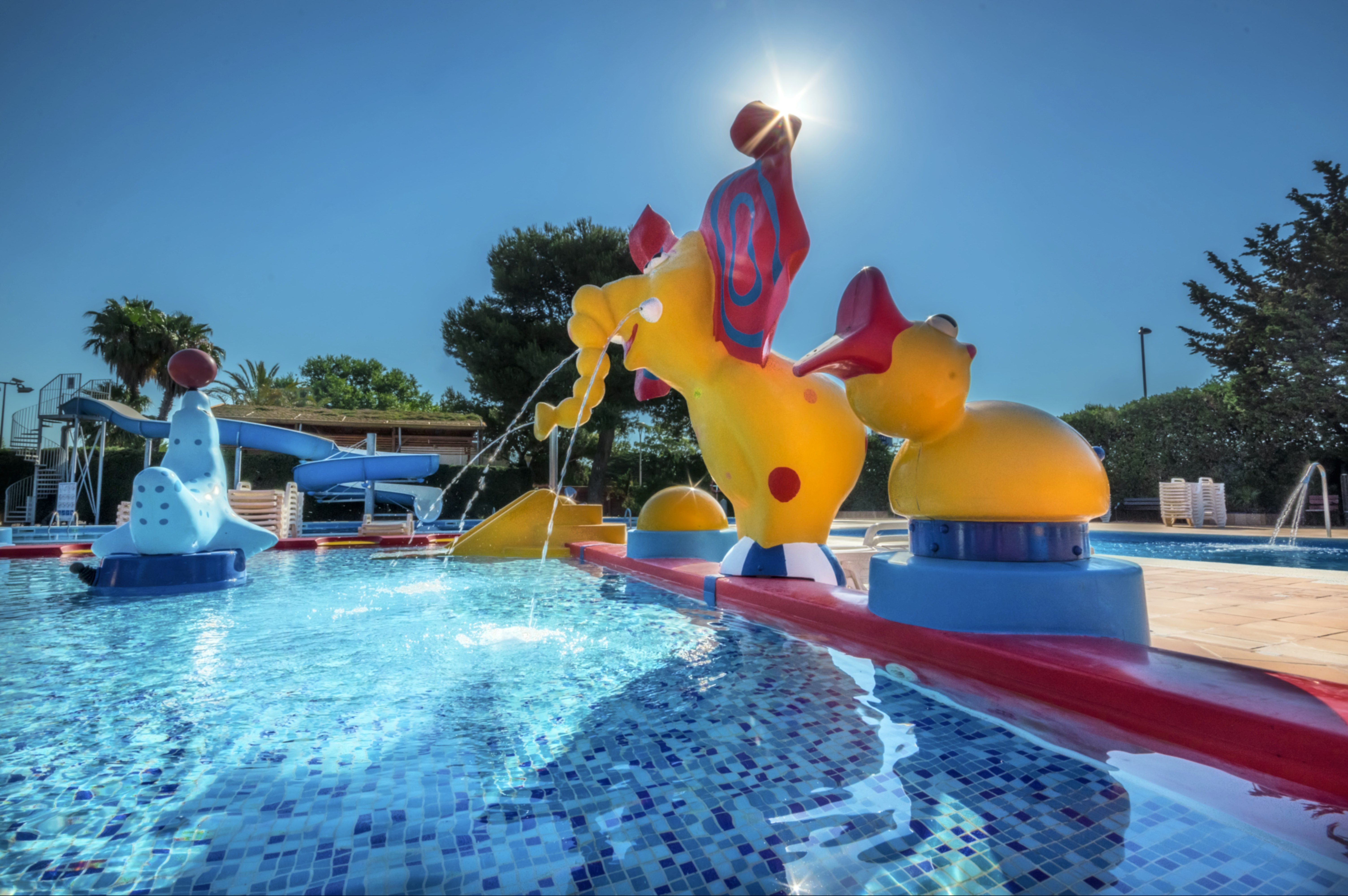 Aufblasfiguren am Wasserspiel im Pool, blaue und gelbe Tiere mit Wasserstrahlen, Sonnenschein und Bäume im Hintergrund.