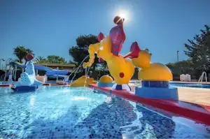 Aufblasfiguren am Wasserspiel im Pool, blaue und gelbe Tiere mit Wasserstrahlen, Sonnenschein und Bäume im Hintergrund.