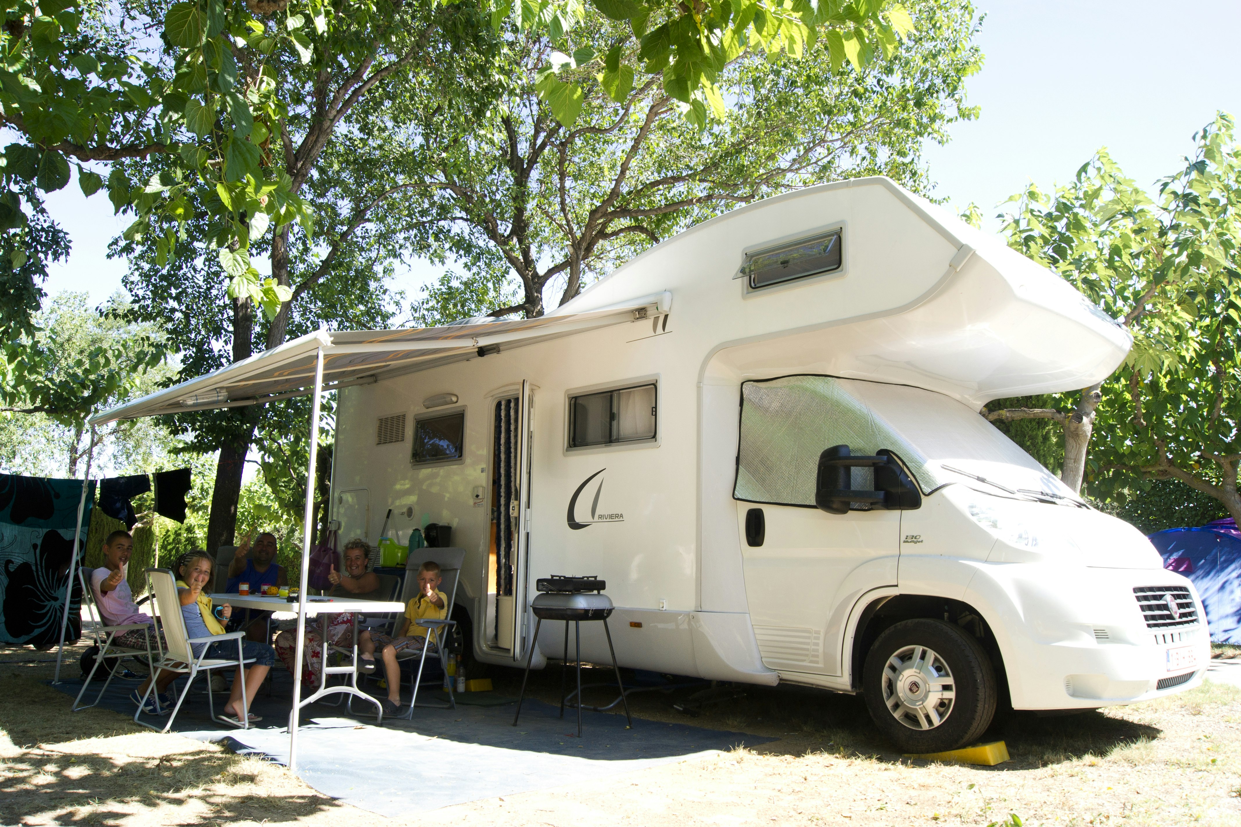 Wohnmobil mit Vorzelt und Sitzgruppe am Campingplatz unter Bäumen, mit Tisch und Stühlen vor dem Fahrzeug.