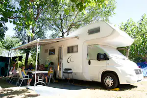 Wohnmobil mit Vorzelt und Sitzgruppe am Campingplatz unter Bäumen, mit Tisch und Stühlen vor dem Fahrzeug.