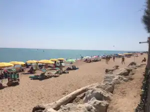 Sandstrand mit Liegestühlen und Sonnenschirmen, im Hintergrund das Meer und ein Weg am Strand mit Personen.