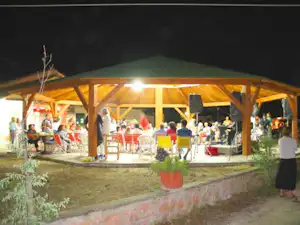 Überdachte Terrasse eines Campingplatz-Restaurants bei Nacht mit beleuchteten Tischen und Stühlen unter Holzpfeilern.