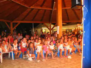 Kinder sitzen dicht beieinander auf weißen Stühlen unter einem überdachten Zelt am Campingplatz.