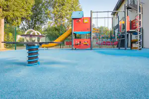 Spielplatz mit Kletterturm und gelber Rutsche, Federwippe auf blauem Boden, Zaun und Bäume im Hintergrund.