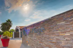Camping-Eingangsschild aus Stein mit beleuchtetem Schriftzug „Cala Plage“ an der Wand, Weg und rote Pflanzkübel.