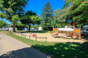 Spielplatz auf dem Campingplatz mit Sandfläche, Holzschaukel und Bänken, umgeben von Bäumen und geparkten Autos.