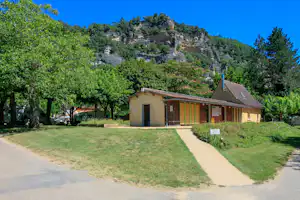 Campingbungalow mit Terrasse am Weg, umgeben von Rasen und Bäumen, im Hintergrund ein bewaldeter Hang.