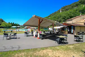 Terrasse am Campingplatz mit Sitzgruppen unter Holzüberdachung, daneben Gebäude, Wege und Blick auf den Poolbereich.