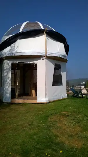 Weißes Kuppelzelt mit offener Veranda auf einer grünen Wiese, Holzpodest und Gartenmöbel im Hintergrund.