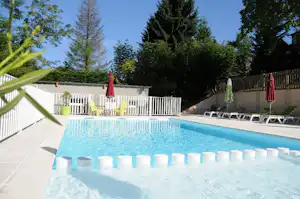 Schwimmbecken eines Campingplatzes mit weißen Liegen am Rand, Zaun und Bäumen im Hintergrund.