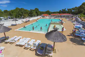 Schwimmbad im Campingbereich mit türkisfarbenem Wasser, Liegen, Sonnenschirmen und Umzäunung, im Hintergrund Gebäude und Bäume.