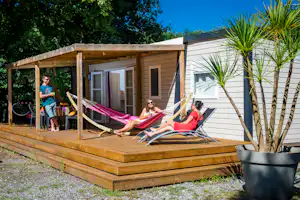 Mobilheim auf Holzterrasse mit Hängematte, Sonnenschirmen und Sitzgelegenheiten, umgeben von Bäumen und grünem Garten.