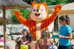 Orangefarbenes Maskottchen mit ausgebreiteten Armen am Pool, Kinder im Vordergrund, Sonnenschirm und Zeltunterkunft im Hintergrund.