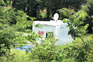 Weißes Wohnmobil mit Heckklappe im Grünen, von Büschen und Bäumen umgeben, Campingplatz mit Rasenfläche im Hintergrund.