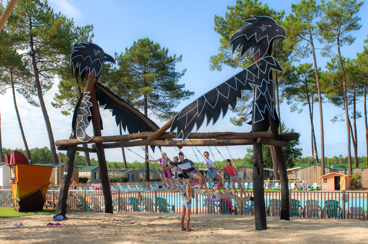 Drachenförmiges Klettergerüst auf dem Sandspielplatz am Campingplatz, Kinder sitzen und stehen am Rand des Pools.