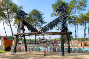 Drachenförmiges Klettergerüst auf dem Sandspielplatz am Campingplatz, Kinder sitzen und stehen am Rand des Pools.
