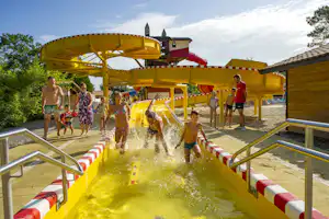 Gelbe Wasserrutsche im Campingbereich, Kinder und Erwachsene rutschen, im Hintergrund Gebäude und Bäume.