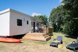Mobilheim mit Holzdeck, Gartenmöbeln und zwei Liegen auf dem Stellplatz, umgeben von Bäumen und Rasen.