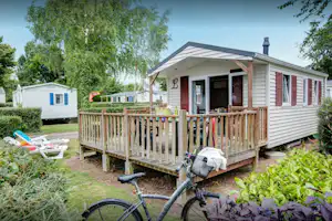 Wohnwagen mit Holzveranda und Stufen, davor ein Fahrrad und Gartenbeete, im Hintergrund weitere Wohnwagen und Bäume.