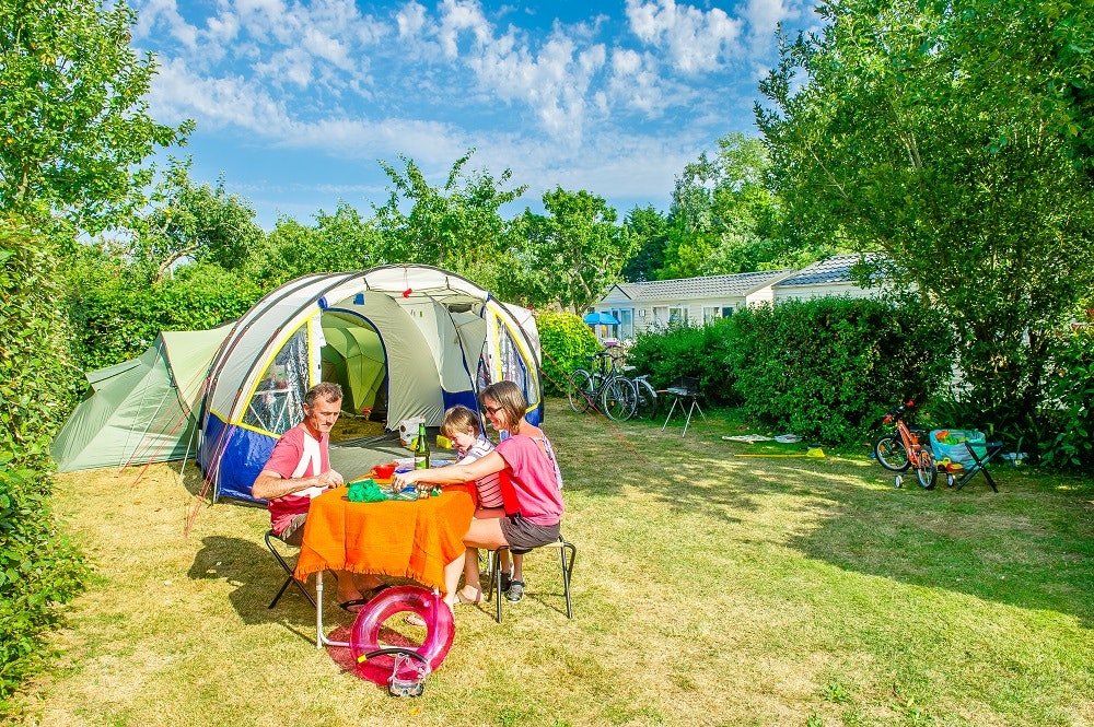 Familienzelt im Campinggarten, Kinder sitzen am Tisch mit buntem Tuch, daneben Spielzeug und Liegestühle im Grünen.