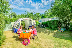 Familienzelt im Campinggarten, Kinder sitzen am Tisch mit buntem Tuch, daneben Spielzeug und Liegestühle im Grünen.
