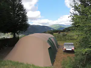 Zelt auf einem Campingplatz am Hang, mit Blick auf Berge, Wiese und einen kleinen Wohnwagen im Hintergrund.