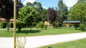 Grüne Campinganlage mit Rasen, Bäumen und Hecken, im Hintergrund Gebäude und ein Kiesweg.