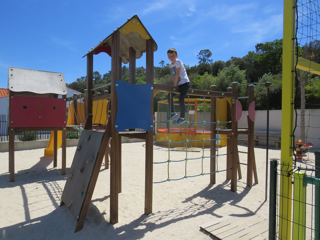 Spielplatz am Campingplatz mit Klettergerüst, Rutsche und Sandboden, im Hintergrund Zaun und Bäume.