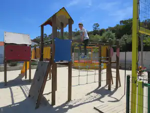 Spielplatz am Campingplatz mit Klettergerüst, Rutsche und Sandboden, im Hintergrund Zaun und Bäume.