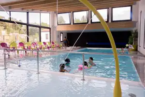 Innenpool mit Glasfront, Liegen und Sonnenschirmen; im Wasser planschen zwei Personen, im Hintergrund Fenster und Pflanzen.