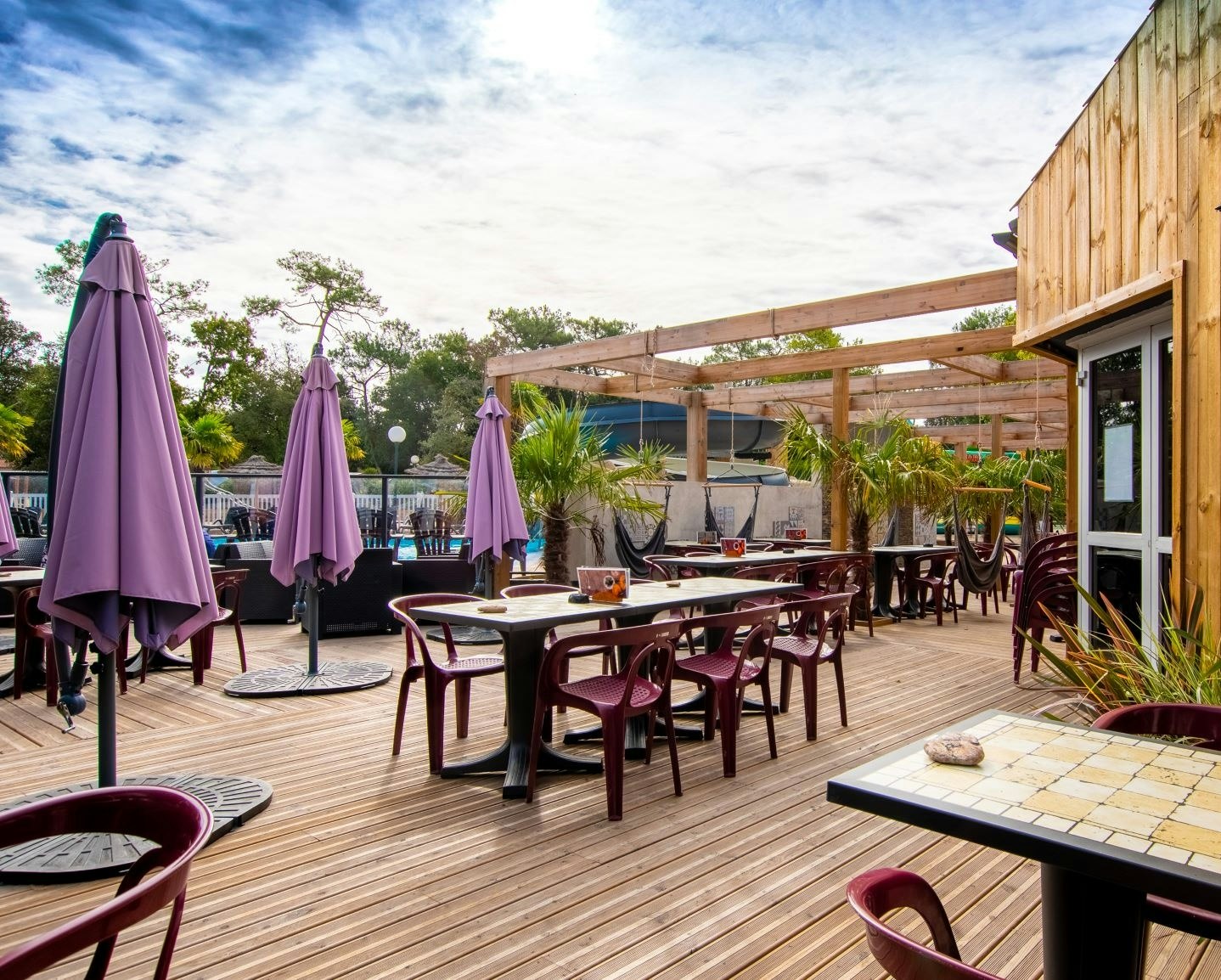 Holzterrasse am Campingplatz mit Tischen, Stühlen und mehreren lila Sonnenschirmen neben einem Holzgebäude.