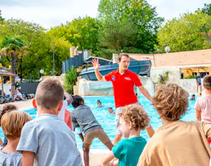 Ein Animateur im roten Shirt zeigt Kindern am Camping-Schwimmbecken eine Bewegung, im Hintergrund Bungalows und Bäume.