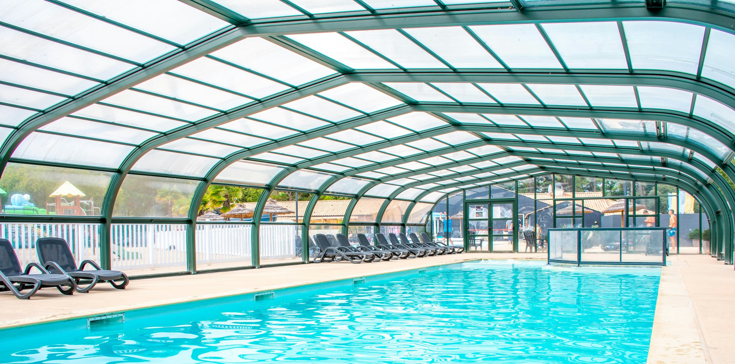 Überdachter Poolbereich mit blauem Becken, Liegen und Glaswänden im Hintergrund.