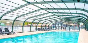 Überdachter Poolbereich mit blauem Becken, Liegen und Glaswänden im Hintergrund.